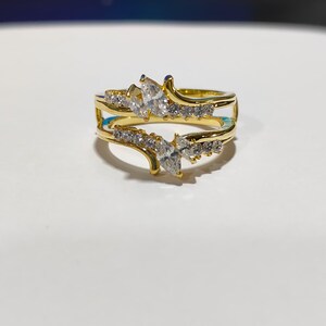 Peut inclure: Une bague dorée avec un design à double bande. La bague présente des pierres claires et scintillantes et des pierres centrales en forme de poire. La bague est posée sur un fond blanc.