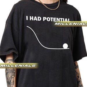 Pode incluir: Camiseta cinza escuro com o texto "I HAD POTENTIAL" acima de um gráfico de linha branca de uma bola rolando ladeira abaixo. A palavra "MILLENNIALS" é impressa em uma faixa dourada na parte inferior da camiseta.