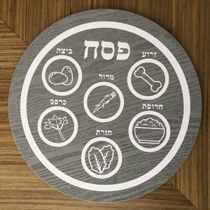 May include: A gray wooden Seder plate with white outlines and Hebrew text. The plate features six sections with symbols for the Passover Seder: matzah, maror, charoset, karpas, beitzah, and bone.