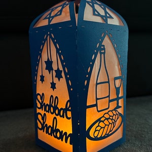 May include: A blue paper lantern with cut-out designs, including a Star of David, a bottle of wine, a glass of wine, a challah bread, and the words "Shabbat Shalom".