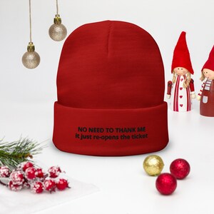 May include: A bright red beanie hat with the text "NO NEED TO THANK ME it just re-opens the ticket" in black. The hat is folded at the brim. Christmas ornaments and decorative figures are in the background.