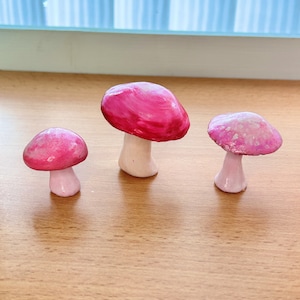 May include: Three pink and white painted ceramic mushrooms of varying sizes. The mushrooms are sitting on a wooden surface.