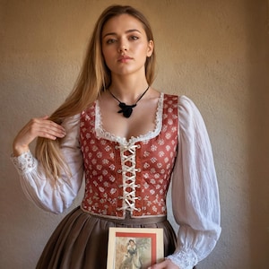 May include: A woman wearing a white blouse with long sleeves and a red and white patterned bodice. She is holding a book with a floral design and a woman in a dress on the cover. The woman is wearing a black necklace with a pendant.