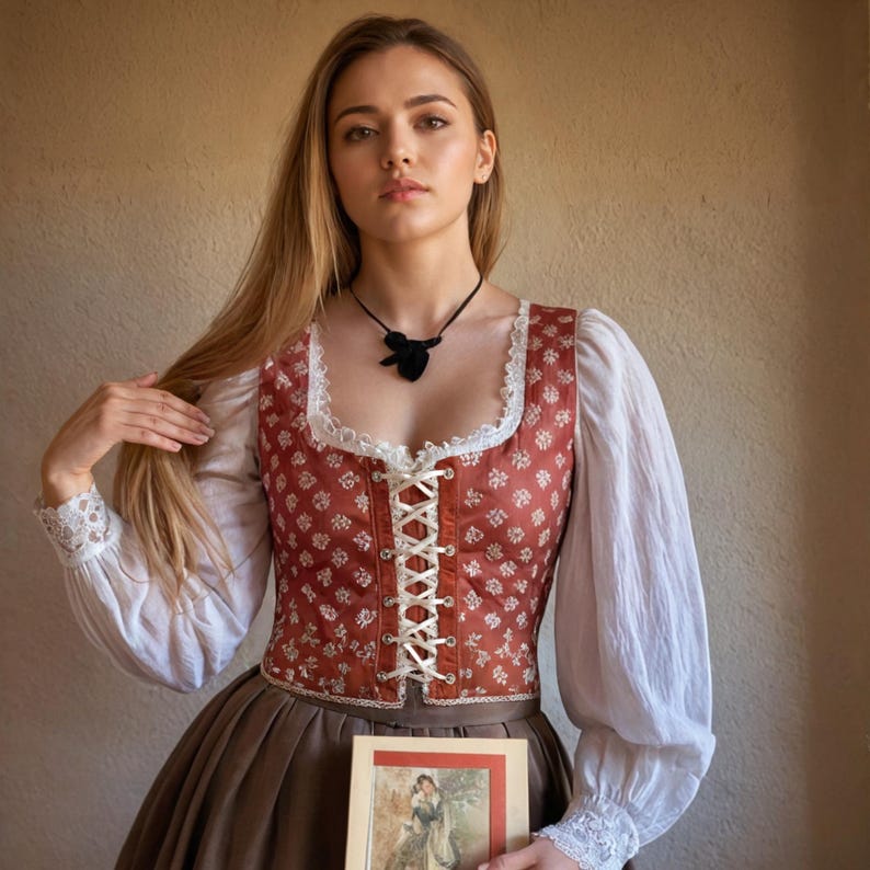 May include: A woman wearing a white blouse with long sleeves and a red and white patterned bodice. She is holding a book with a floral design and a woman in a dress on the cover. The woman is wearing a black necklace with a pendant.