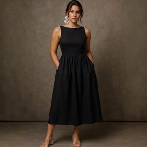 May include: A sleeveless black dress with a boat neckline and a gathered waist. The dress has pockets and falls to mid-calf. The model is wearing clear heels and earrings. The background is a solid brown.