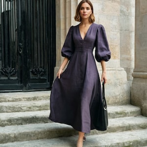 May include: A woman wearing a dark purple linen dress with puffed sleeves and a V-neckline. She is holding a black leather handbag.