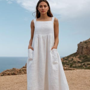 May include: A white linen dress with a square neckline and wide straps. The dress has a gathered waist and two large pockets. The model is standing outdoors with the ocean and sky in the background.