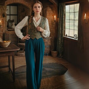 May include: A woman wearing a white blouse with a green and gold patterned vest and dark blue wide-leg pants. She is standing in a room with a wooden floor and a window in the background.
