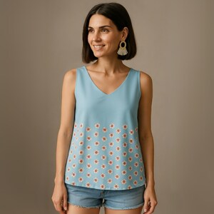 May include: A sleeveless, light blue top with a V-neck. The bottom half of the top features a repeating pattern of small white flowers with orange centers. The top is paired with denim shorts.