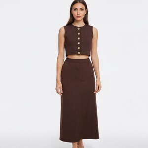 May include: A two-piece brown outfit featuring a sleeveless crop top with gold buttons and a matching high-waisted midi skirt. The outfit is made of a solid, smooth fabric and is suitable for various occasions.