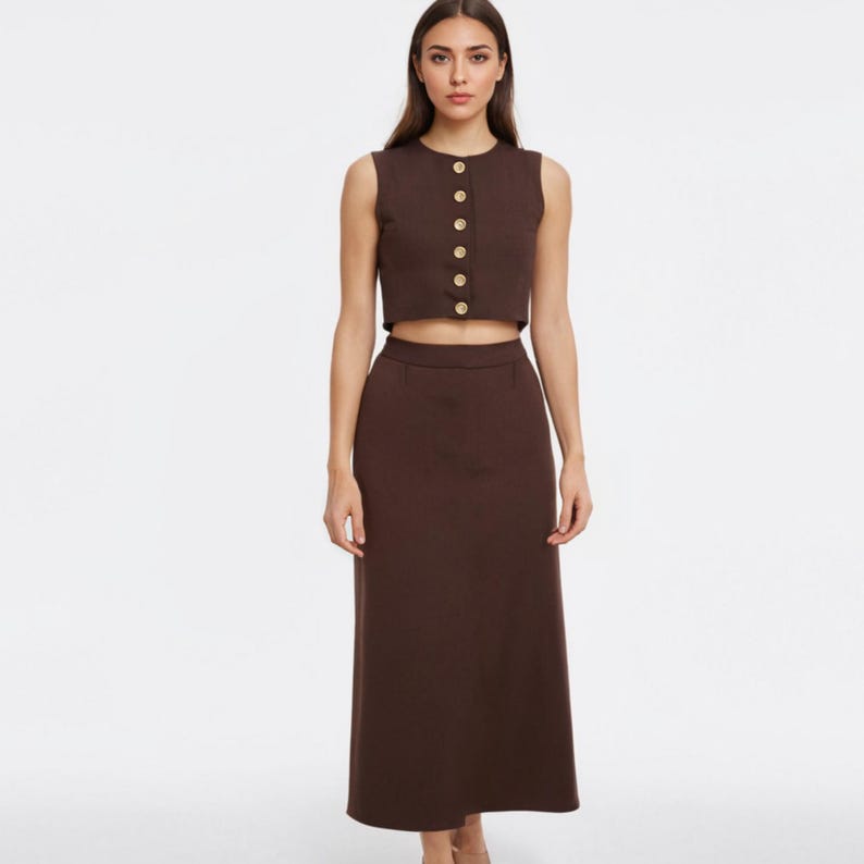 May include: A two-piece brown outfit featuring a sleeveless crop top with gold buttons and a matching high-waisted midi skirt. The outfit is made of a solid, smooth fabric and is suitable for various occasions.