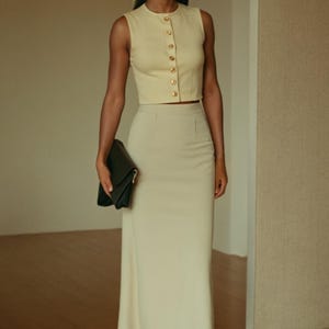 May include: A woman wearing a sleeveless, cream-colored top with gold buttons and a matching cream-colored skirt. She is holding a black clutch.