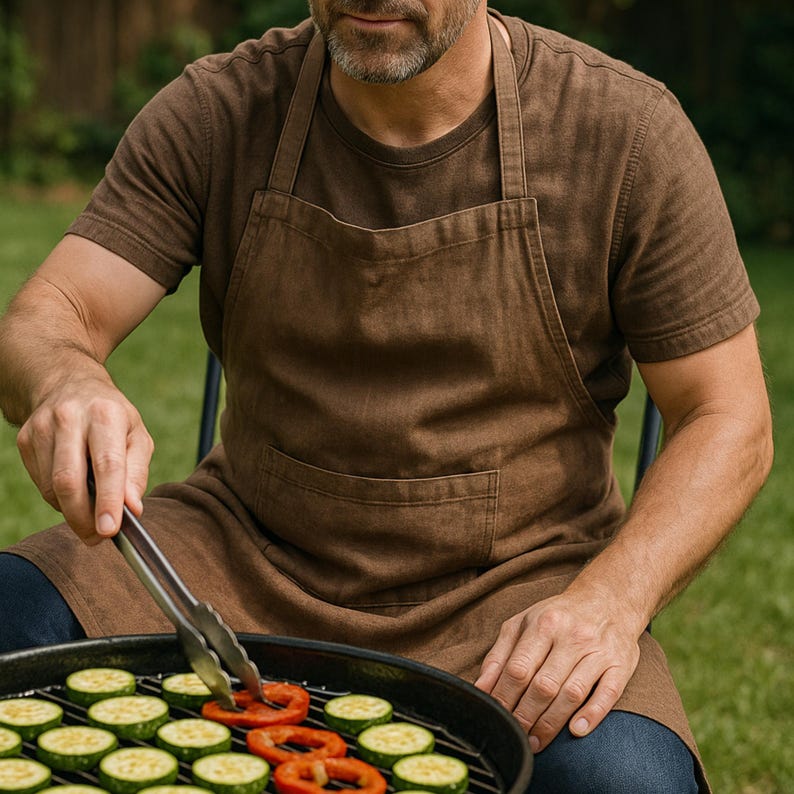 May include: A brown apron with a front pocket is worn by a person grilling vegetables on a barbecue. The person is wearing a brown t-shirt and using tongs to turn the food. Sliced zucchini and red bell peppers are visible on the grill.