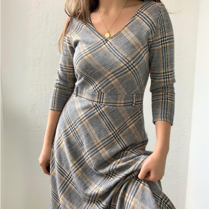 May include: A long-sleeved dress with a plaid pattern in shades of gray and yellow. The dress has a v-neckline and a fitted bodice. The skirt is full and flows to the ankles.