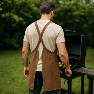 May include: A brown apron with crisscross straps is worn over a light-colored t-shirt. The apron is made of a textured material. The person is standing near a black grill, suggesting outdoor cooking and grilling.