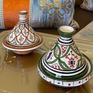 May include: Two ceramic tagine pots with conical lids. One is white with brown and orange patterns, the other is green with red and blue floral designs. The pots sit on a gold surface, with decorative cushions in the background.