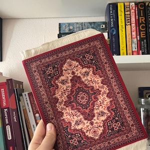 May include: A small, rectangular rug featuring a detailed floral design in red, cream, and navy blue. The rug has a cream-coloured fringe along the edges. This is likely a decorative item or a miniature rug.