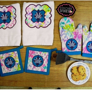May include: A set of kitchen towels, pot holders, oven mitts, and a spatula, all featuring a floral pattern with a monogrammed "ME" in a blue circle. The set is on a wooden surface with a plate of cookies.