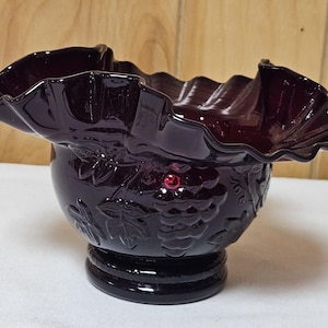 May include: A deep purple glass bowl featuring a wide, ruffled rim and a molded exterior pattern of grape clusters and leaves. The bowl sits on a circular pedestal base, with the dark glass reflecting light across its textured, embossed surface.