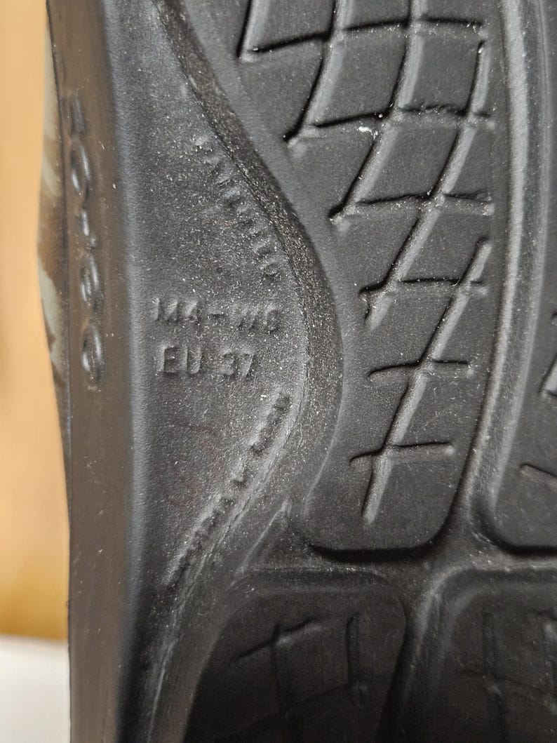 May include: Close-up of the sole of a black shoe. The sole has a textured pattern with diamond shapes and cross-shaped grooves. The size is marked as 144-W9, EU 37.