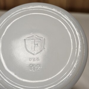 Pu&ograve; includere: Primo piano del fondo di un oggetto in ceramica bianca. La base presenta un design ad anelli concentrici in rilievo. In rilievo al centro, uno scudo con la lettera "F", la parola "USA" e il numero "112".