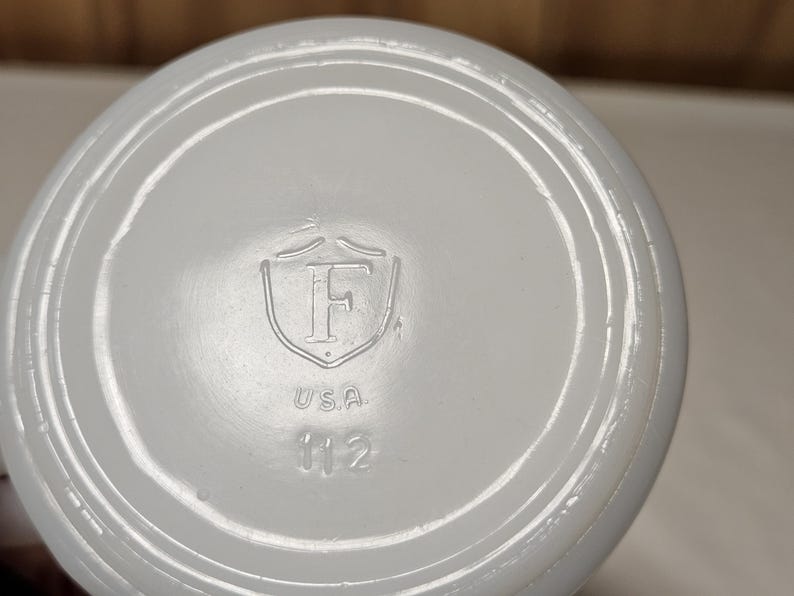 Pu&ograve; includere: Primo piano del fondo di un oggetto in ceramica bianca. La base presenta un design ad anelli concentrici in rilievo. In rilievo al centro, uno scudo con la lettera "F", la parola "USA" e il numero "112".