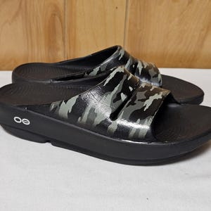 May include: Pair of black slide sandals with a camouflage pattern on the straps. The sandals have a thick sole and a contoured footbed. The brand logo is visible on the side of the sole.