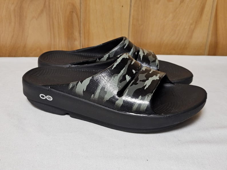 May include: Pair of black slide sandals with a camouflage pattern on the straps. The sandals have a thick sole and a contoured footbed. The brand logo is visible on the side of the sole.