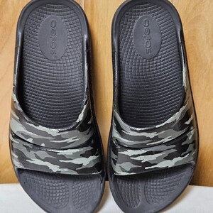 May include: A pair of black slide sandals with a camouflage pattern in shades of gray and green. The sandals have a textured footbed and the brand name "OOFOS" is visible on the insole.