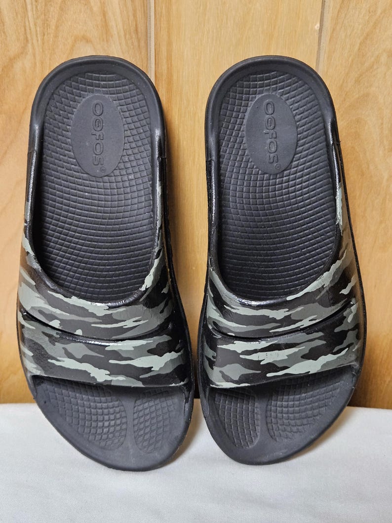 May include: A pair of black slide sandals with a camouflage pattern in shades of gray and green. The sandals have a textured footbed and the brand name "OOFOS" is visible on the insole.