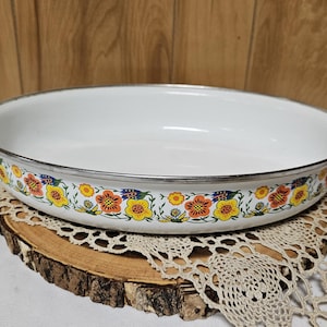 May include: White enamel oval baking dish with a floral design in yellow, orange, and blue. The dish has a silver rim and white handles. It is displayed on a wooden slice and lace doily.