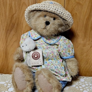 May include: A plush teddy bear wearing a floral dress with blue trim and a woven hat. The bear is holding a small white stuffed animal and a tag that reads "Gen-yoo-wine BOYDS". The bear's fur is light brown.