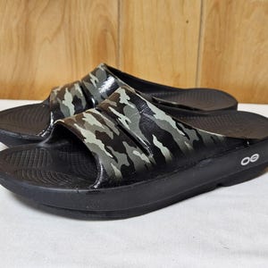 May include: A pair of black slide sandals with a camouflage pattern on the straps. The sandals have a thick sole and the brand logo is visible on the side. The sandals are designed for comfort and casual wear.