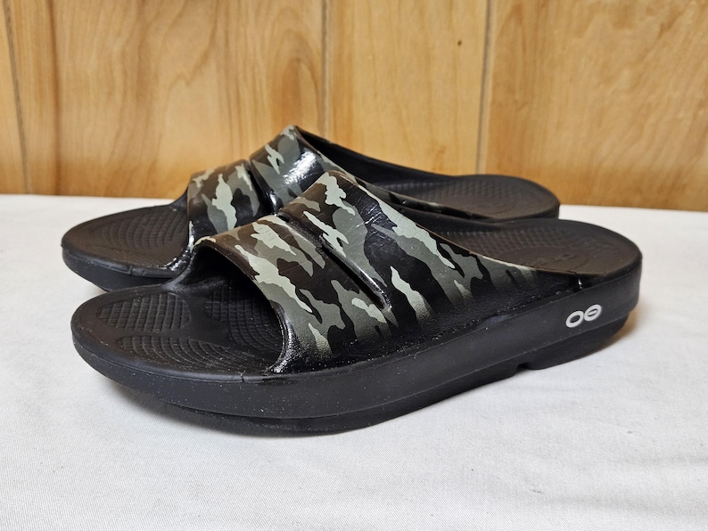 May include: A pair of black slide sandals with a camouflage pattern on the straps. The sandals have a thick sole and the brand logo is visible on the side. The sandals are designed for comfort and casual wear.