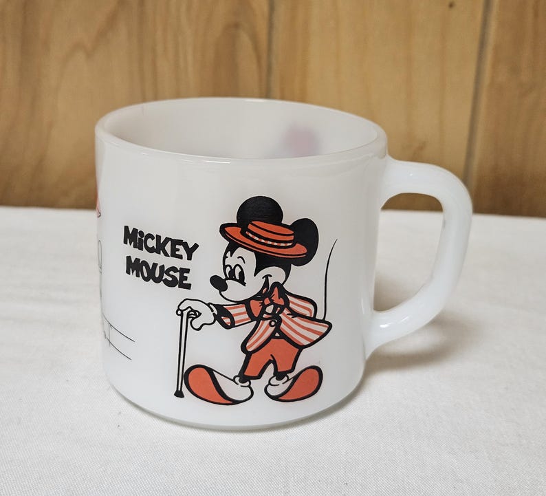 Pu&ograve; includere: Tazza in ceramica bianca con Topolino in un completo rosso e arancione con un bastone. La tazza ha un manico curvo e le parole "Mickey Mouse" stampate sul lato. Il design &egrave; in stile vintage.