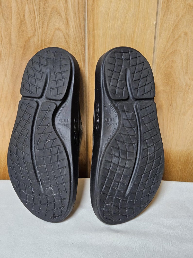 May include: A pair of black sandals, viewed from the soles. The sandals have a textured, patterned design on the soles. The sandals are made of a solid, dark material. The sandals are EU size 37.