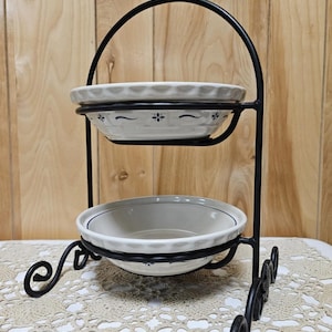 May include: Two-tiered serving stand with white ceramic bowls and a black wrought iron frame. The bowls have a scalloped edge and blue floral accents. The stand has decorative scrollwork on the base and handle.