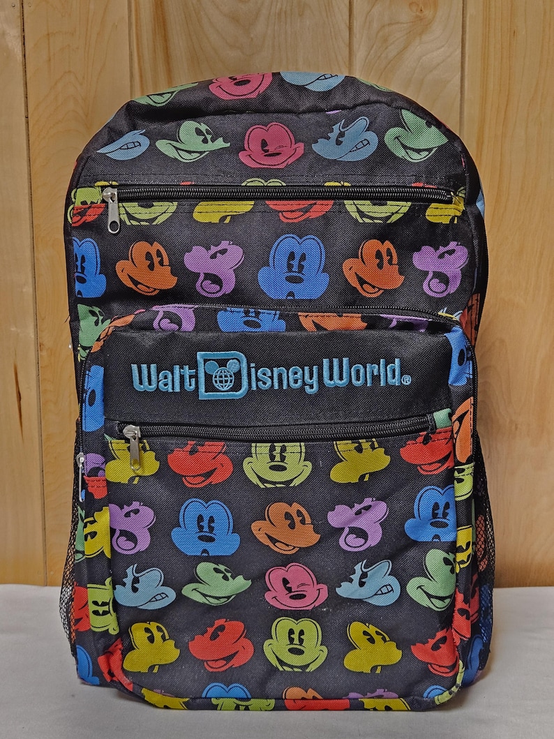 Puede incluir: Una mochila negra con un patr&oacute;n repetitivo de siluetas de cabezas de Mickey Mouse coloridas en rosa, verde, azul, naranja y amarillo. El frente incluye un bolsillo con cremallera con el texto bordado Walt Disney World en azul claro.