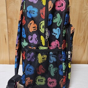 Puede incluir: Una mochila negra con un patr&oacute;n repetitivo de siluetas de cabezas de Mickey Mouse coloridas en tonos azul, verde, naranja, morado, rojo y amarillo. La bolsa incluye un bolsillo lateral de malla, un compartimento frontal con cremallera y correas negras.