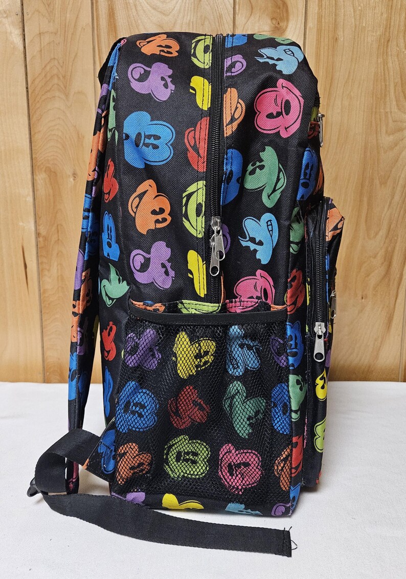 Puede incluir: Una mochila negra con un patr&oacute;n repetitivo de siluetas de cabezas de Mickey Mouse coloridas en tonos azul, verde, naranja, morado, rojo y amarillo. La bolsa incluye un bolsillo lateral de malla, un compartimento frontal con cremallera y correas negras.