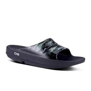 May include: Black slide sandal with a camouflage pattern across the top. The sandal has a thick sole and the brand logo is visible on the side. The camouflage design features shades of green and gray.