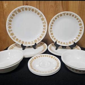 May include: A collection of white Corelle dinnerware with a gold floral pattern. The set includes dinner plates, salad plates, saucers, and bowls. The plates are displayed on black plate stands.