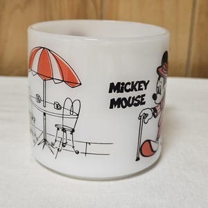 Pu&ograve; includere: Tazza in ceramica bianca con un'illustrazione nera e rossa di Topolino in un'ambientazione da caff&egrave;. Il design include un ombrellone a strisce arancioni e bianche, un tavolo e le parole "Mickey Mouse".