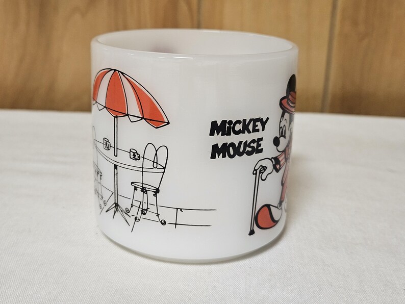 Pu&ograve; includere: Tazza in ceramica bianca con un'illustrazione nera e rossa di Topolino in un'ambientazione da caff&egrave;. Il design include un ombrellone a strisce arancioni e bianche, un tavolo e le parole "Mickey Mouse".