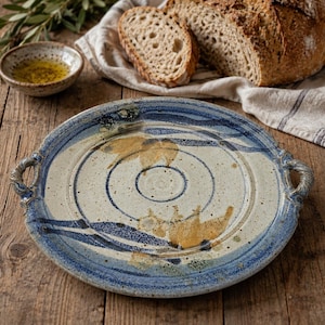 May include: A round ceramic serving platter with two side handles, featuring a speckled cream base with blue and golden-brown abstract painted accents. The platter has a textured surface with concentric circular indentations and a rustic finish.