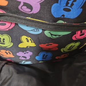 Puede incluir: Una mochila negra con un patr&oacute;n repetitivo de cabezas de rat&oacute;n de dibujos animados coloridas en tonos azul, verde, naranja, rosa y morado. La bolsa incluye un bolsillo frontal con cremallera y un compartimento principal con forro interior negro.