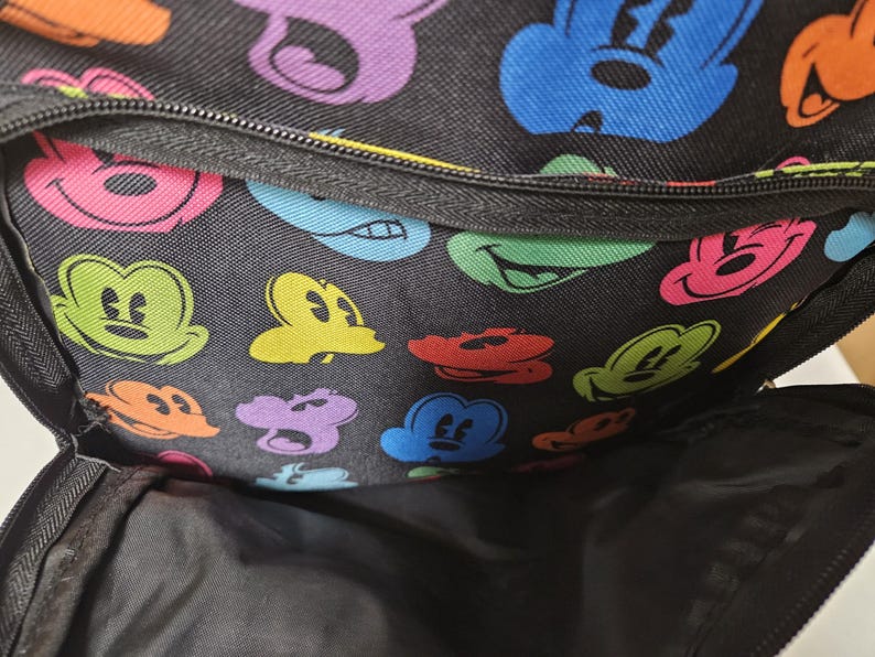 Puede incluir: Una mochila negra con un patr&oacute;n repetitivo de cabezas de rat&oacute;n de dibujos animados coloridas en tonos azul, verde, naranja, rosa y morado. La bolsa incluye un bolsillo frontal con cremallera y un compartimento principal con forro interior negro.