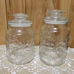 May include: Two clear glass jars with lids. The jars have a textured surface with a fruit and leaf design. The jars are of different sizes and sit on a cream-colored lace doily.