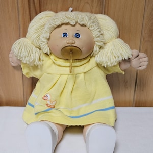 May include: A vintage doll with blonde yarn hair styled in pigtails. The doll has blue eyes and a pacifier. It wears a yellow dress with a duck graphic and blue stripes. White shoes complete the outfit.