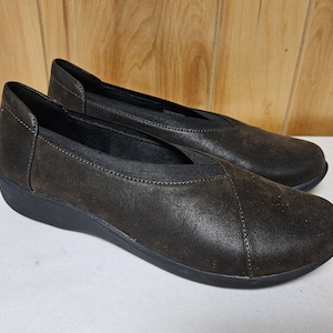 May include: A pair of dark brown slip-on shoes featuring a textured finish, a rounded toe, and a black elastic gore panel at the instep. The shoes have a low-profile black sole and visible stitching along the front and sides of the upper section.